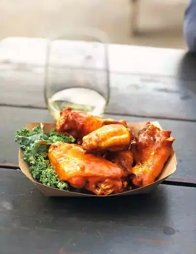 A to go container with chicken wings, kale garnish, and a glass of white wine on a picnic table